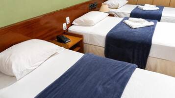 Habitación de hotel con tres camas individuales, cada una con ropa blanca y una manta azul. Cuenta con un cabecero de madera, una mesita de noche con lámpara y un teléfono.