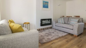 Ett vardagsrum med en grå soffa, en mindre grå fåtölj, en liten träpall, en inramad tavla på väggen och en TV som är monterad på väggen.
