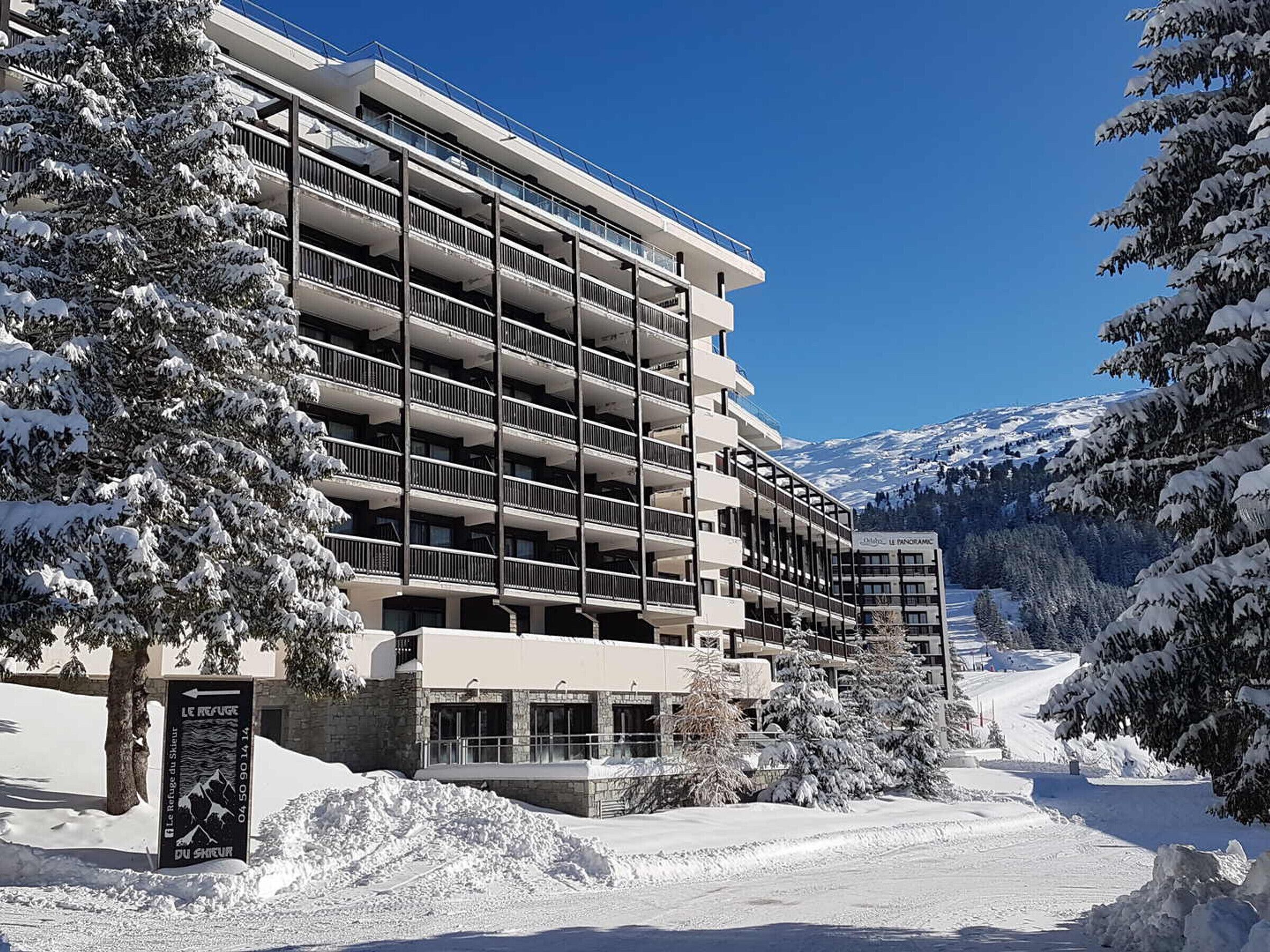 A multi-story hotel with balconies, surrounded by snow-covered trees and mountains.