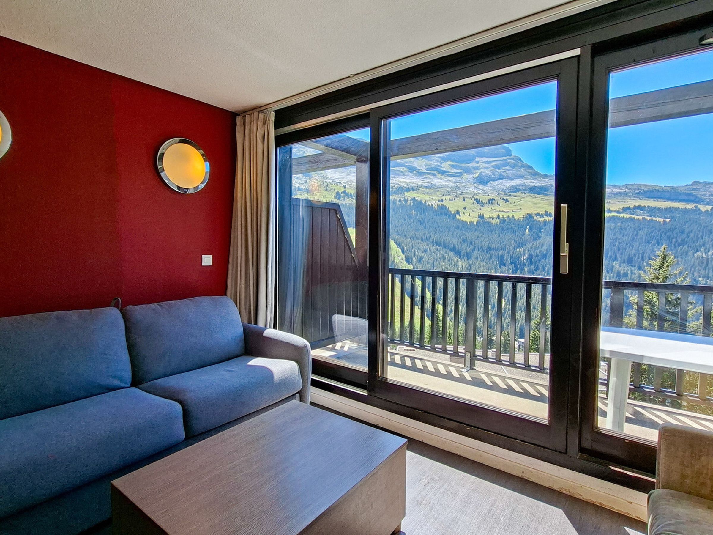 Una sala de estar moderna con un sofá y una mesa de centro, una pared destacada en rojo y un gran ventanal corredero que da a un balcón con vistas a la montaña.