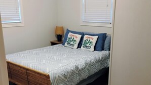 Ein Schlafzimmer mit einem Bett, zwei Kissen, einem Nachttisch, einer Lampe und zwei Fenstern.