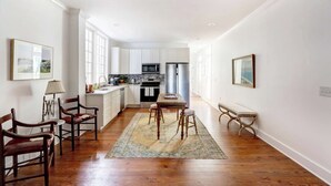 A spacious kitchen with white cabinets, a central island, and a dining area with wooden chairs and a rug. - The Jones Street Parlor | Historic Retreat (Savannah)