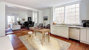 A spacious living area with a dining table, chairs, a kitchen with a large window, and a view of the outside. - The Jones Street Parlor | Historic Retreat (Savannah)
