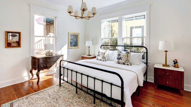 A bedroom with a bed, bedside tables, a chandelier, and large windows. - The Jones Street Parlor | Historic Retreat (Savannah)