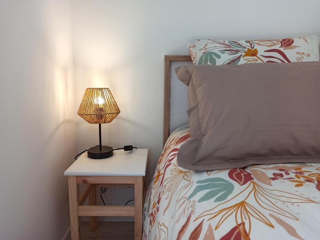 Una cama con una colcha de estampado floral y una sola almohada marrón, una mesita de noche con una lámpara y una pared lisa.