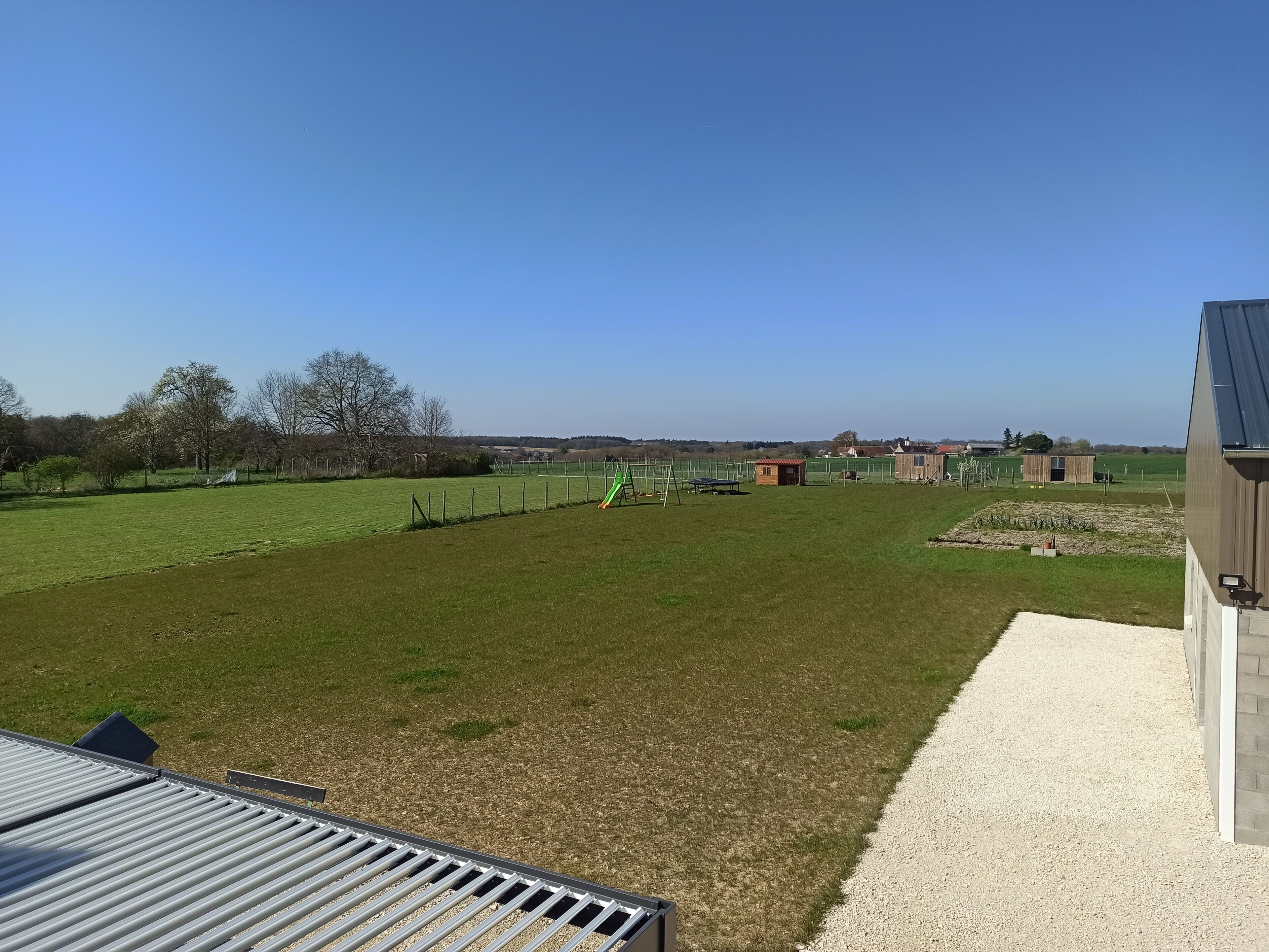 Un campo cubierto de césped con un parque infantil, un edificio pequeño y un granero a lo lejos.