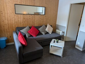 Ein Zimmer mit einem grauen Sofa, einem kleinen weißen Beistelltisch, einer blauen Mülltonne und einer Holzwand mit einem Spiegel.