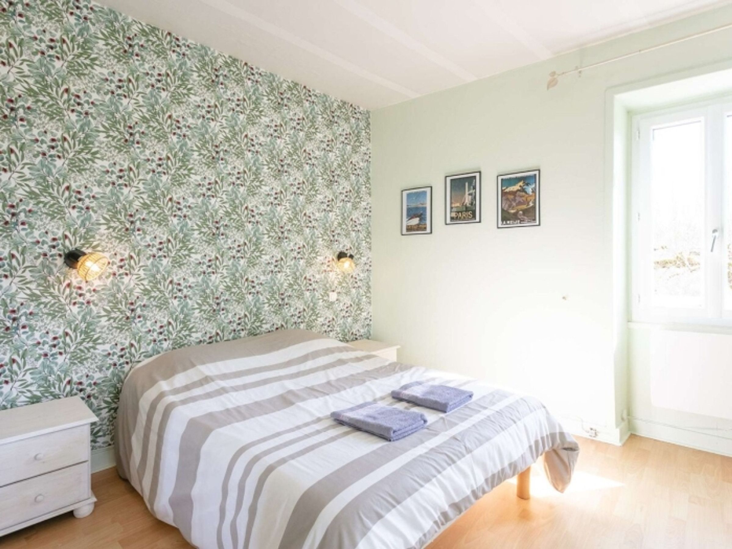 A bedroom with a bed, a nightstand, and a wall with a leafy pattern. There are three framed pictures on the wall.