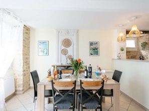 A dining area with a wooden table set for four, surrounded by chairs. There is a fireplace with decorative items and a vase with flowers. The room has a stone wall and a window with curtains. - House in bastide, pets allowed - 5 guests (MONTFAUCON)