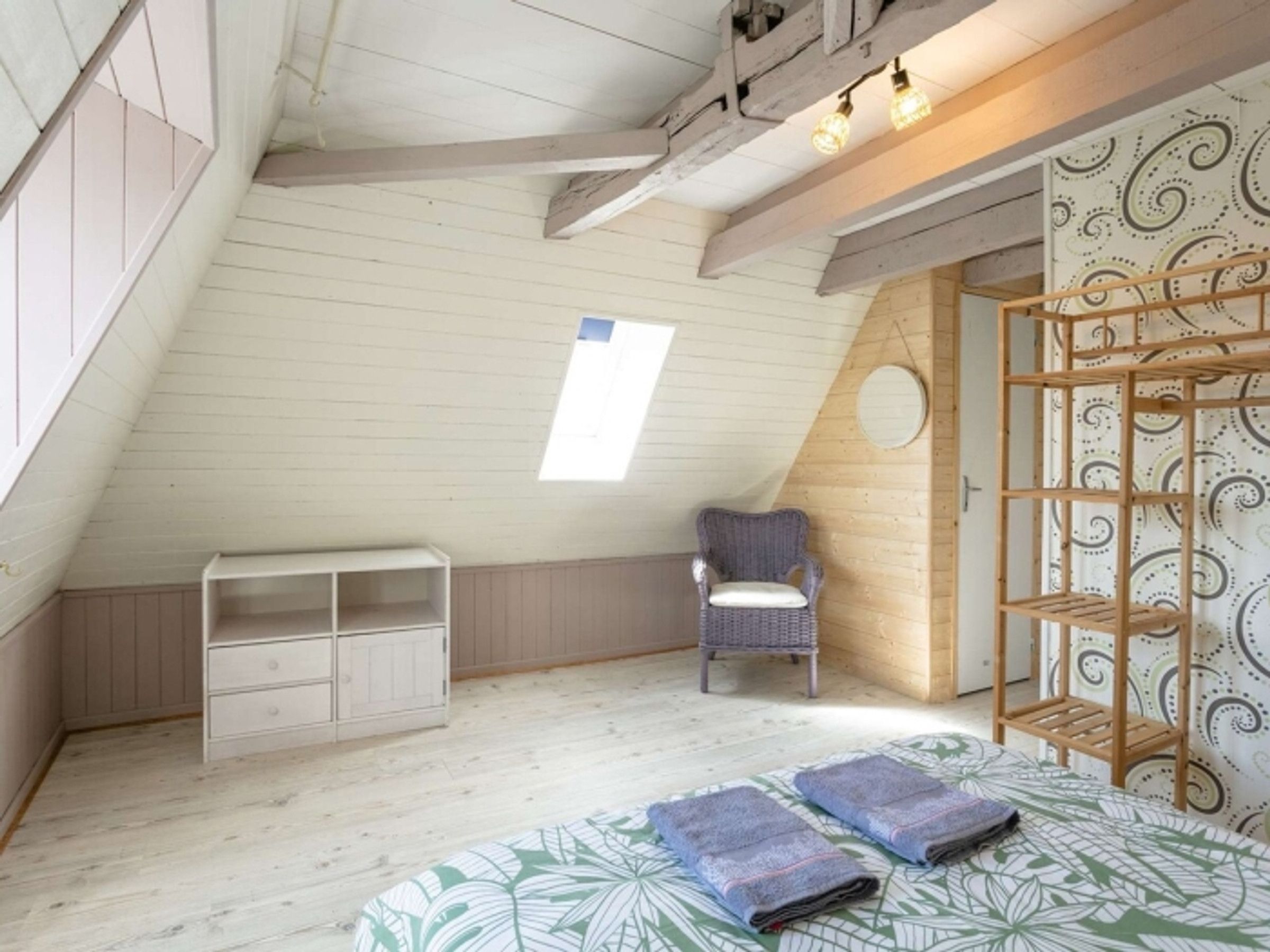 A room with a sloped ceiling, a built-in bench, a chair, a wooden shelf unit, and a bed with a patterned bedspread.