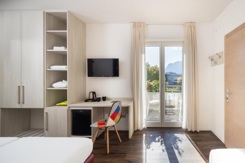 A hotel room with a bed, a desk with a chair, a TV, a small fridge, and a balcony with a view.