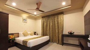 A hotel room with a bed, bedside table, curtains, a desk, and a ceiling fan. - Casa In Luxury Suites (Thiruvananthapuram)