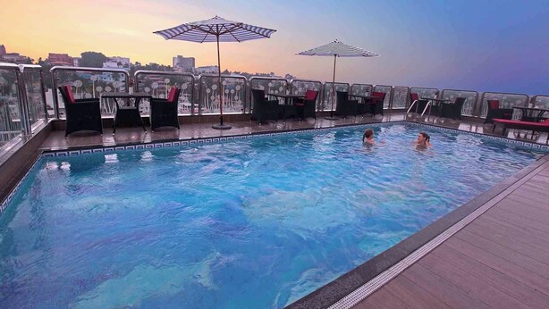 A rooftop pool with clear blue water, surrounded by glass railings, and two umbrellas providing shade. - Casa In Luxury Suites (Thiruvananthapuram)