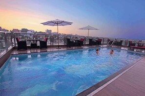 A rooftop pool with clear blue water, surrounded by glass railings, and two umbrellas providing shade.