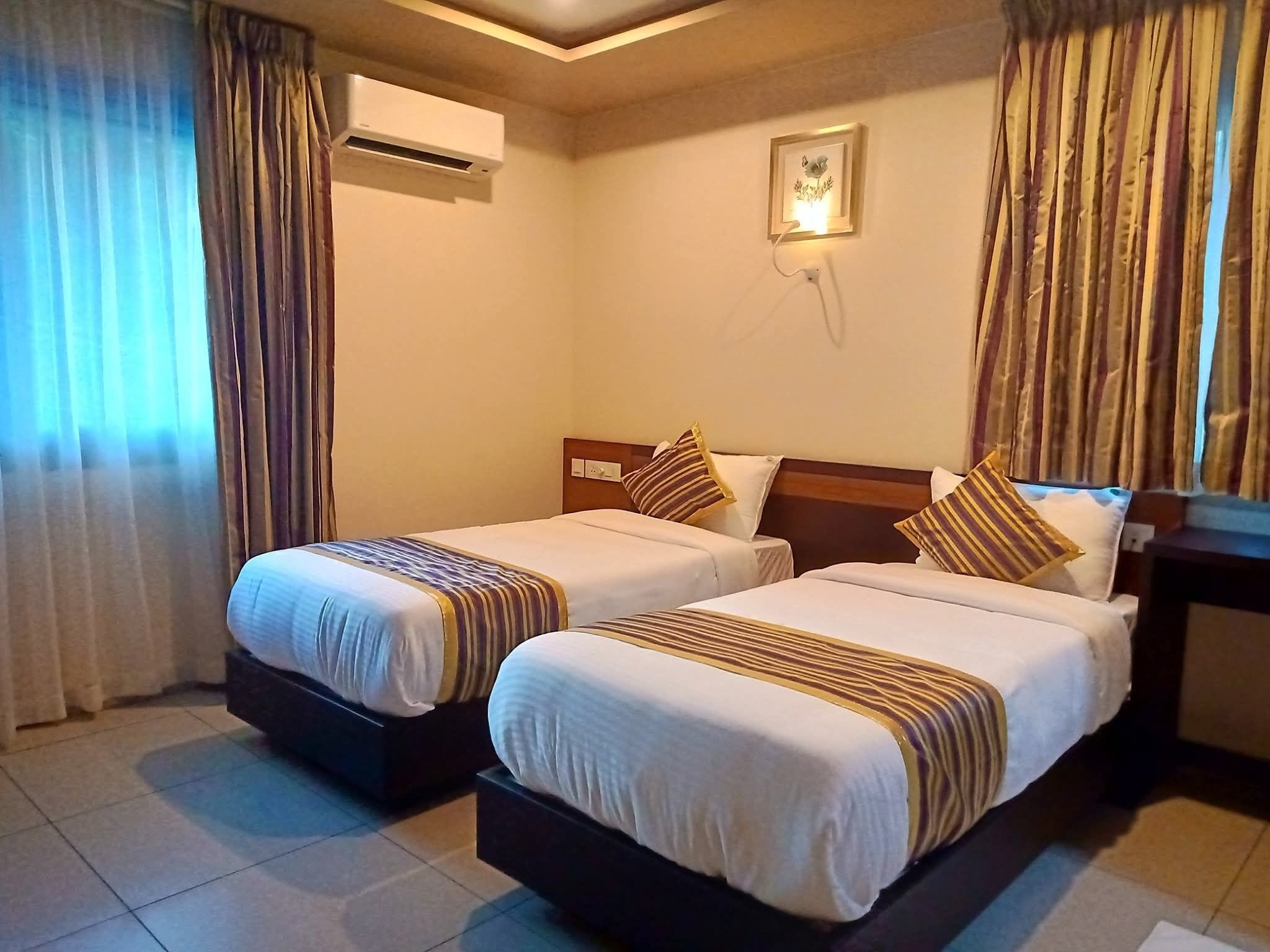 A hotel room with two beds, a nightstand, an air conditioning unit, and a wall-mounted light fixture.