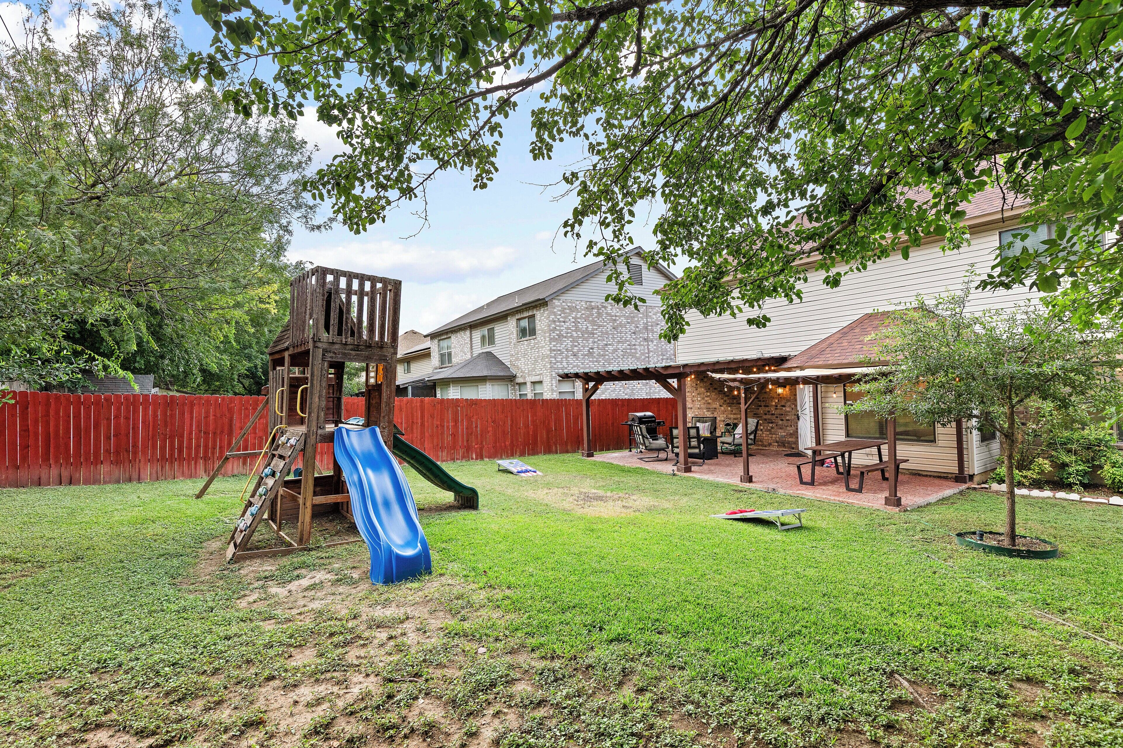 A backyard with a wooden playset, a blue slide, a brick patio, and a covered seating area.