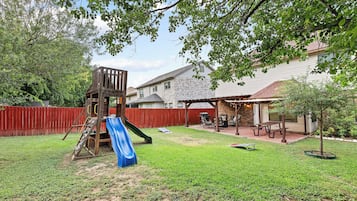 A backyard with a wooden playset, a blue slide, a brick patio, and a covered seating area.