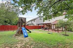 Ein Garten mit einer Holzspieleinrichtung, einer blauen Rutsche, einer Ziegelterrasse und einem überdachten Sitzbereich.