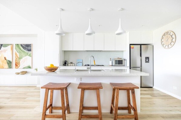 A modern kitchen with white cabinets, a large island with stools, pendant lights, and a clock on the wall.