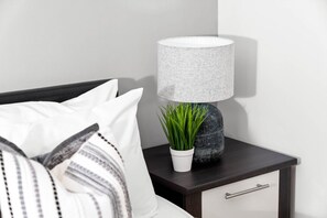 A neatly made bed with white and striped pillows, a dark wooden nightstand with a textured lamp, a small white pot with green plants, and a dark-colored drawer. - City Centre Apartment - Balcony - On Street Parking - Smart Tv & Neflix (Birmingham)