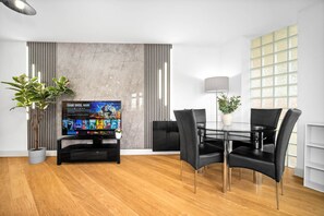 A modern living room with a flat-screen TV, a dining table with chairs, a potted plant, and a large window with blinds. - City Centre Apartment - Balcony - On Street Parking - Smart Tv & Neflix (Birmingham)