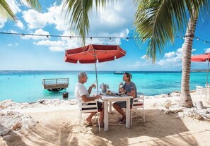 Una parella dinant a una taula al costat de la platja, amb un parasol vermell, vista a l'oceà i un petit moll al fons.