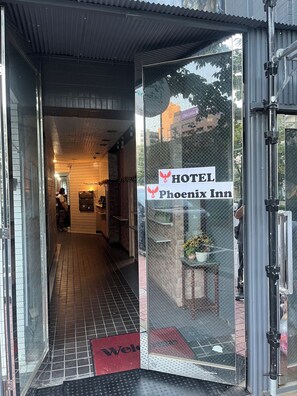 L’entrée d’un hôtel, avec un paillasson où est écrit « Bienvenue », une enseigne indiquant « Hôtel Phoenix Inn », et un couloir menant à l’intérieur.