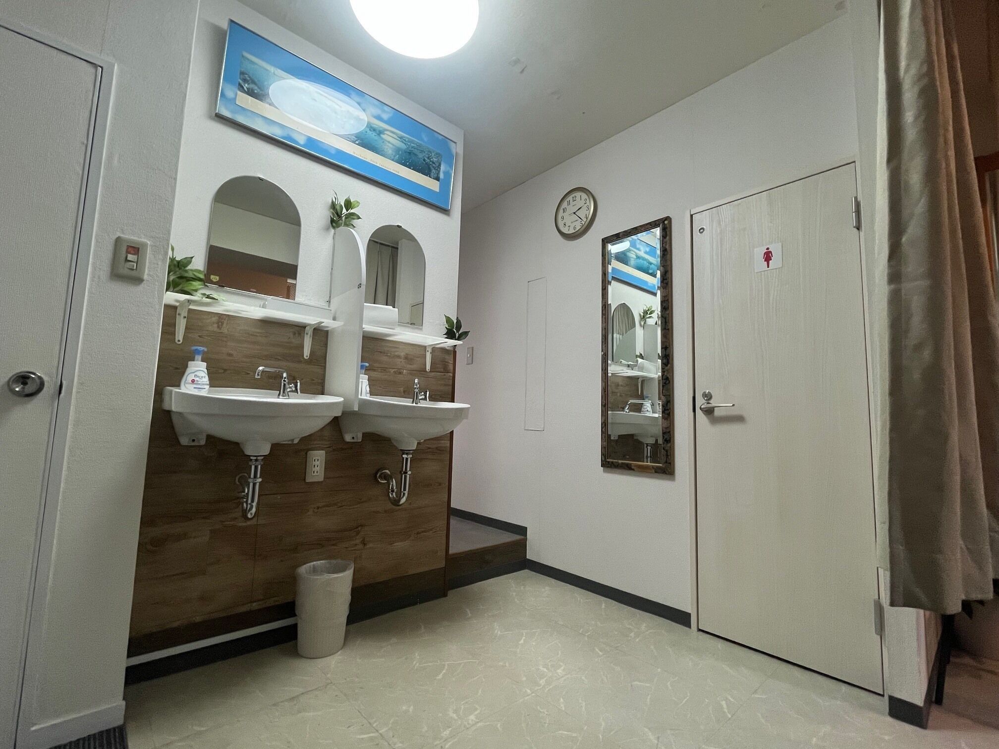 A bathroom with two sinks, a mirror, a clock, and a door with a female restroom symbol.