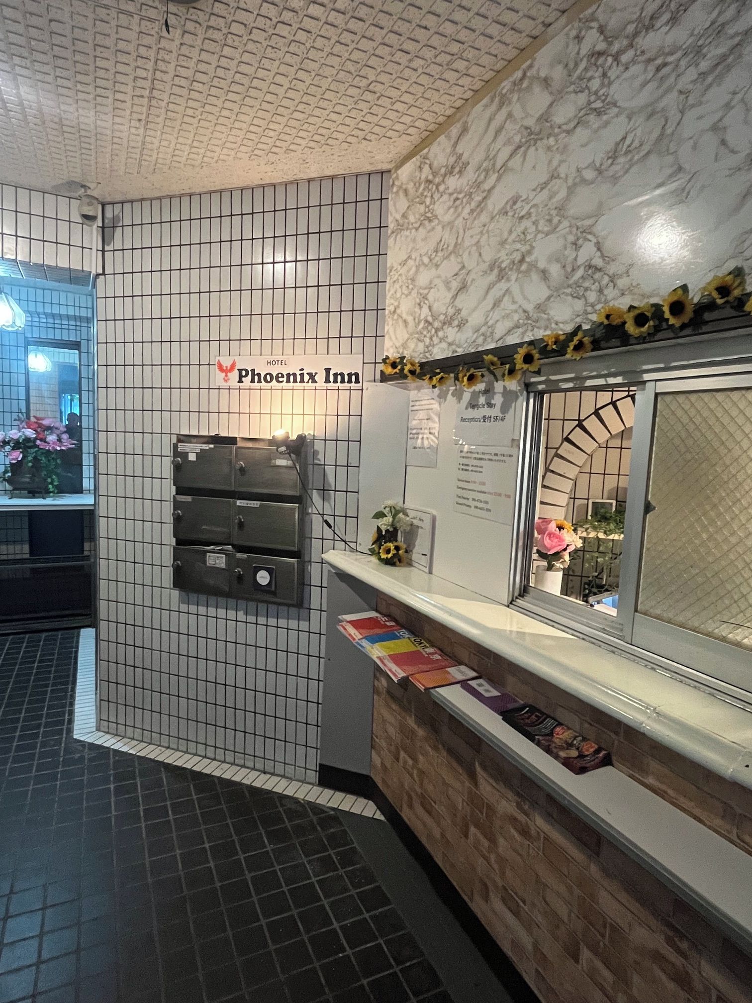 Uma área de recepção com placa do Phoenix Inn, parede azulejada, detalhe em mármore e balcão com folhetos informativos.