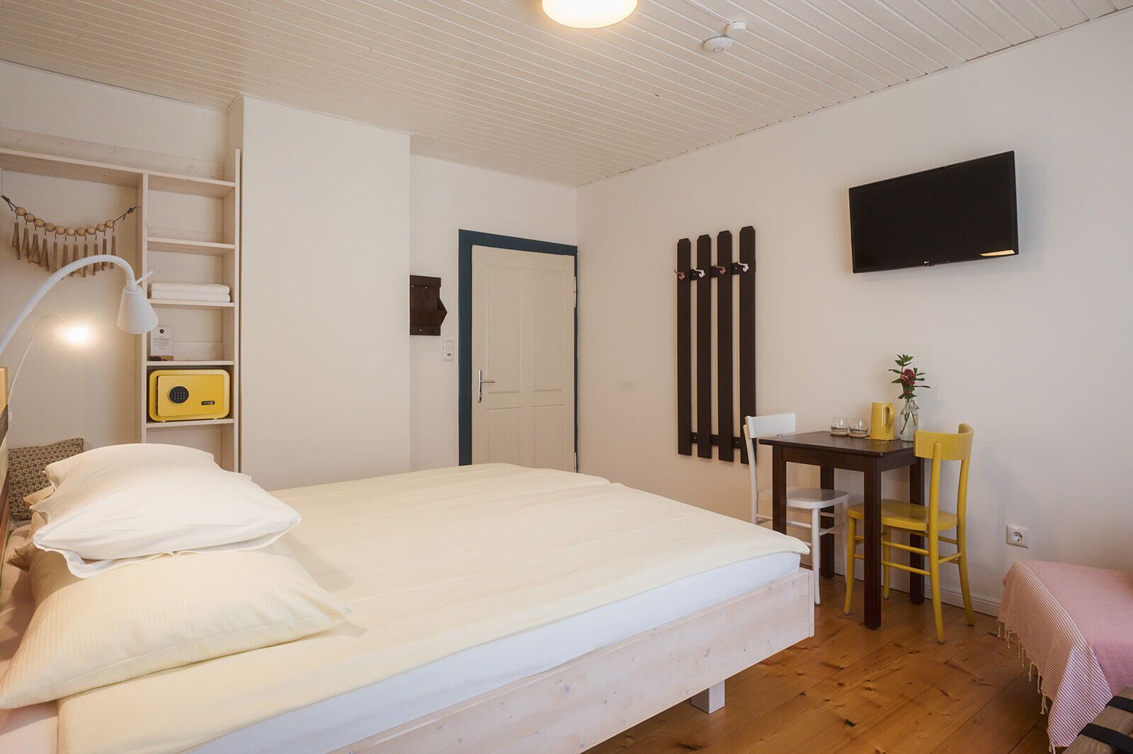 Un dormitorio con cama, lámpara de noche, estante con radio, escritorio con silla y televisor montado en la pared.