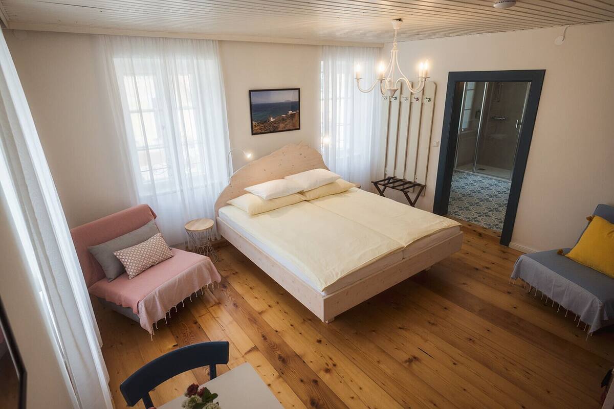 A bedroom with a large bed, a chair, a small table with flowers, a chandelier, and a view of the sea through a window.