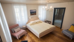 Un dormitorio con una cama grande, una silla, una mesita con flores, un candelabro y vista al mar a través de una ventana.