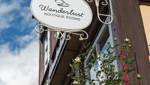 Letrero de 'Wanderlust Boutique Rooms' con logotipo de montaña, sujeto a un edificio con balcón y plantas trepadoras.