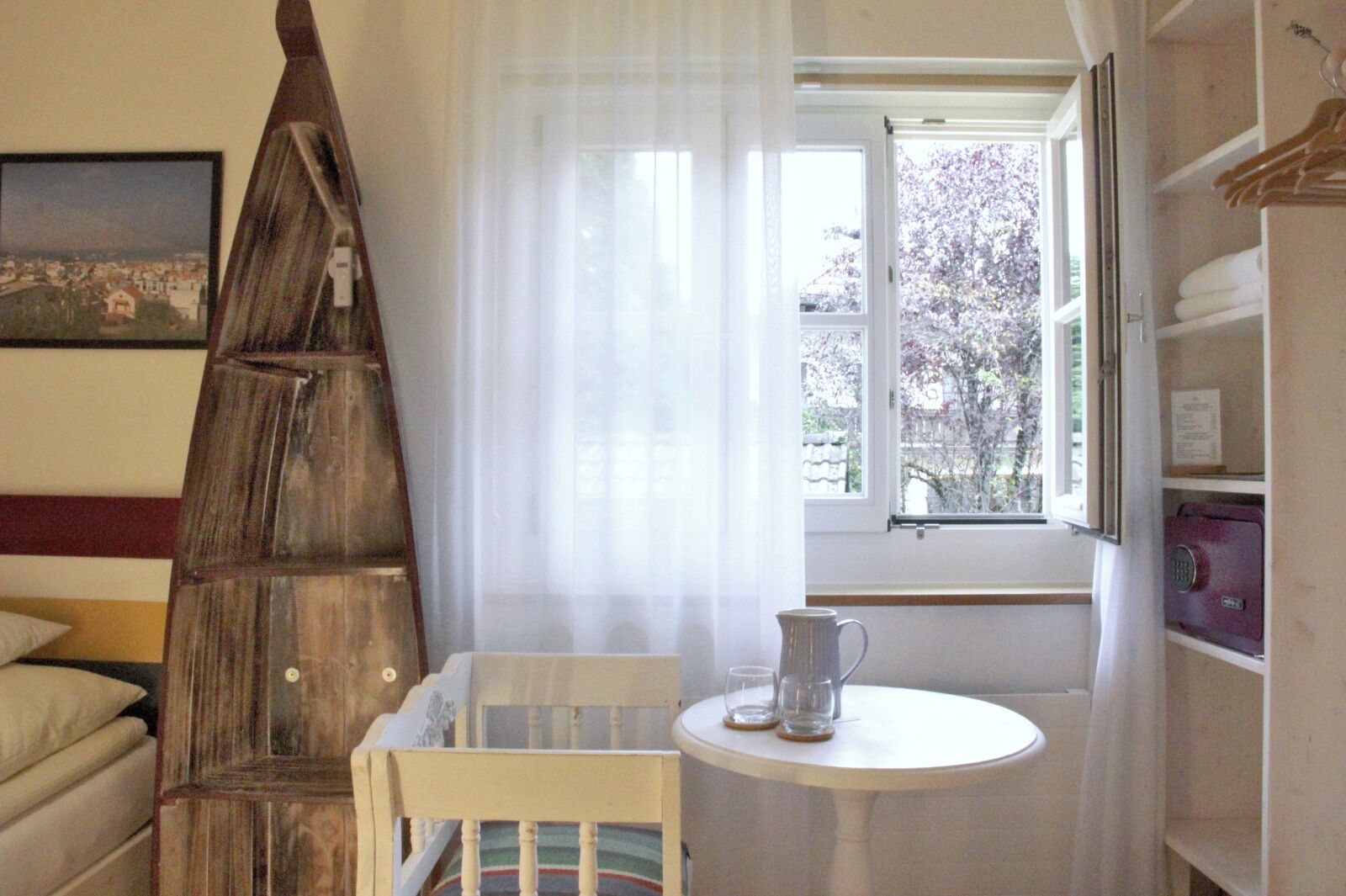 A room with a wooden ladder, a small table with a pitcher, a chair, a window with a view of trees, and a bed with a pillow.