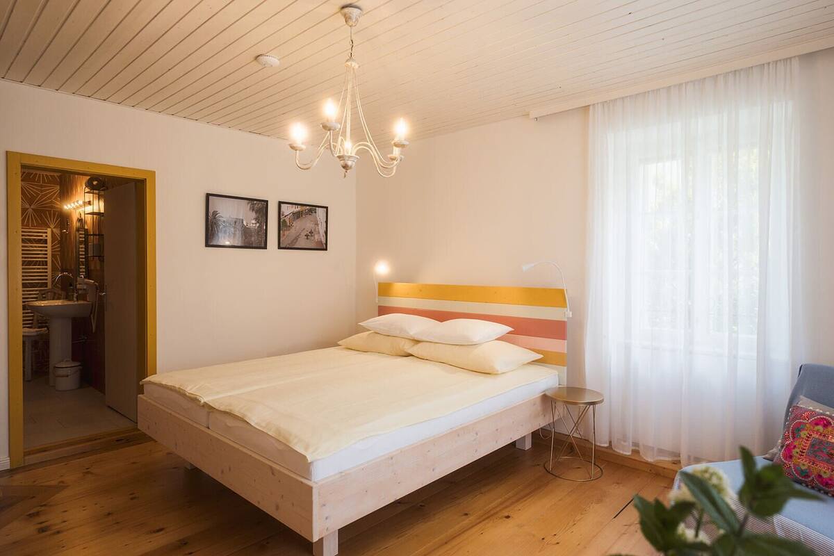A bedroom with a wooden bed, a chandelier, a bedside table, and a window with sheer curtains.