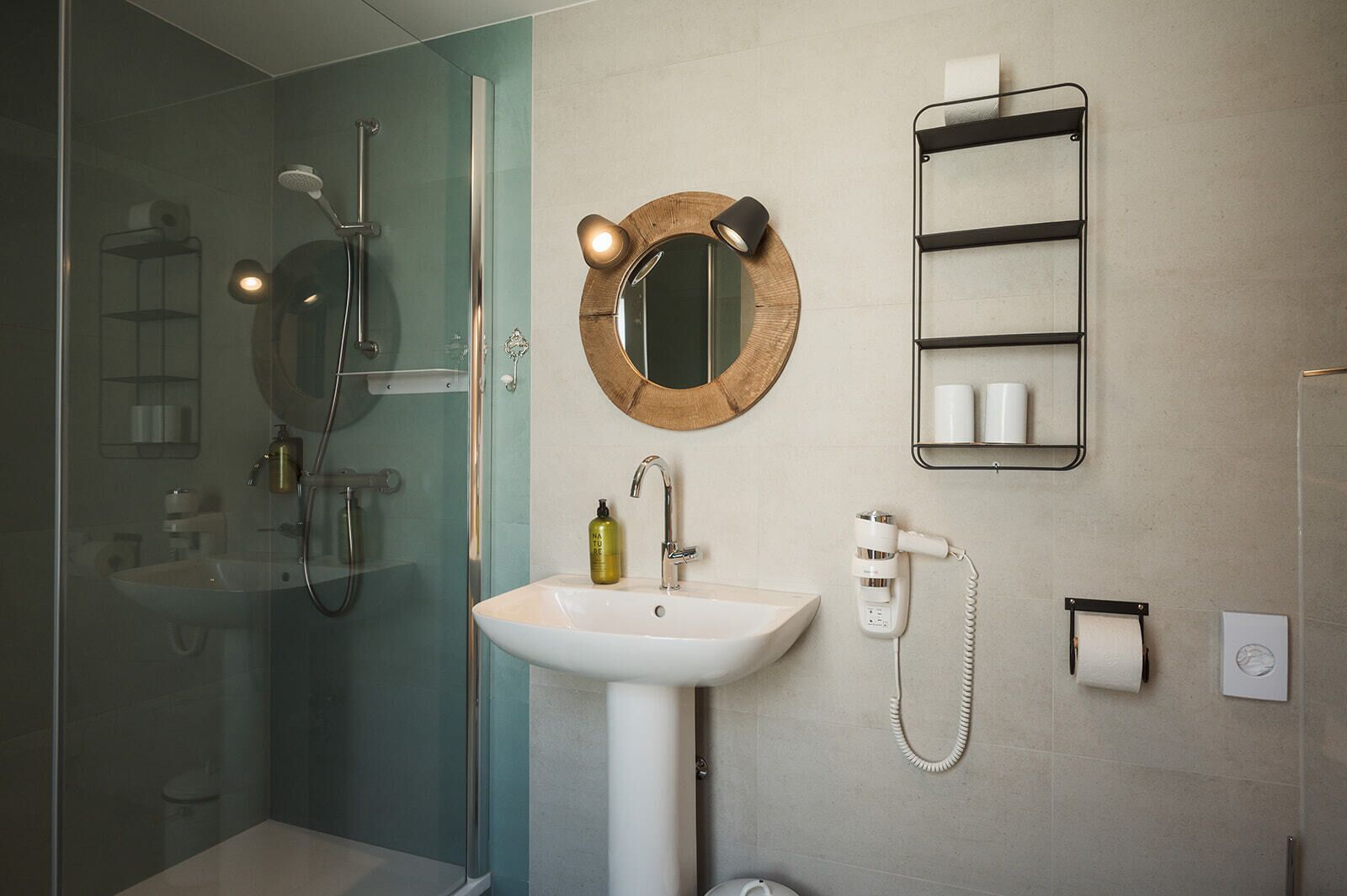 A modern bathroom with a glass shower enclosure, a white sink, a round mirror, a towel rack, and a hairdryer.