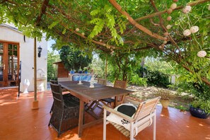 Un patio al aire libre con mesa y sillas de madera, rodeado de exuberante vegetación y una pérgola.