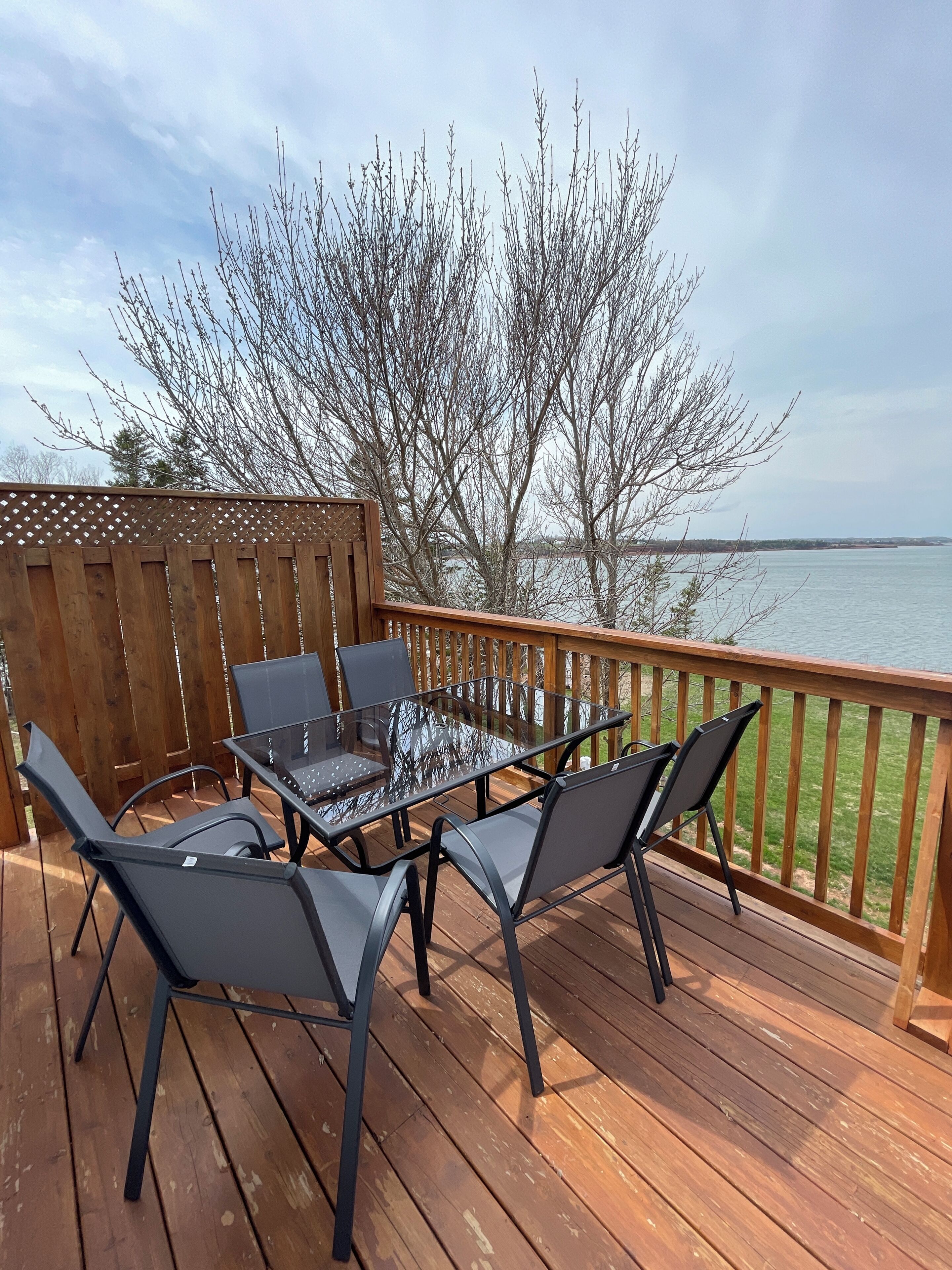 A wooden deck with a glass table and chairs, overlooking a body of water.