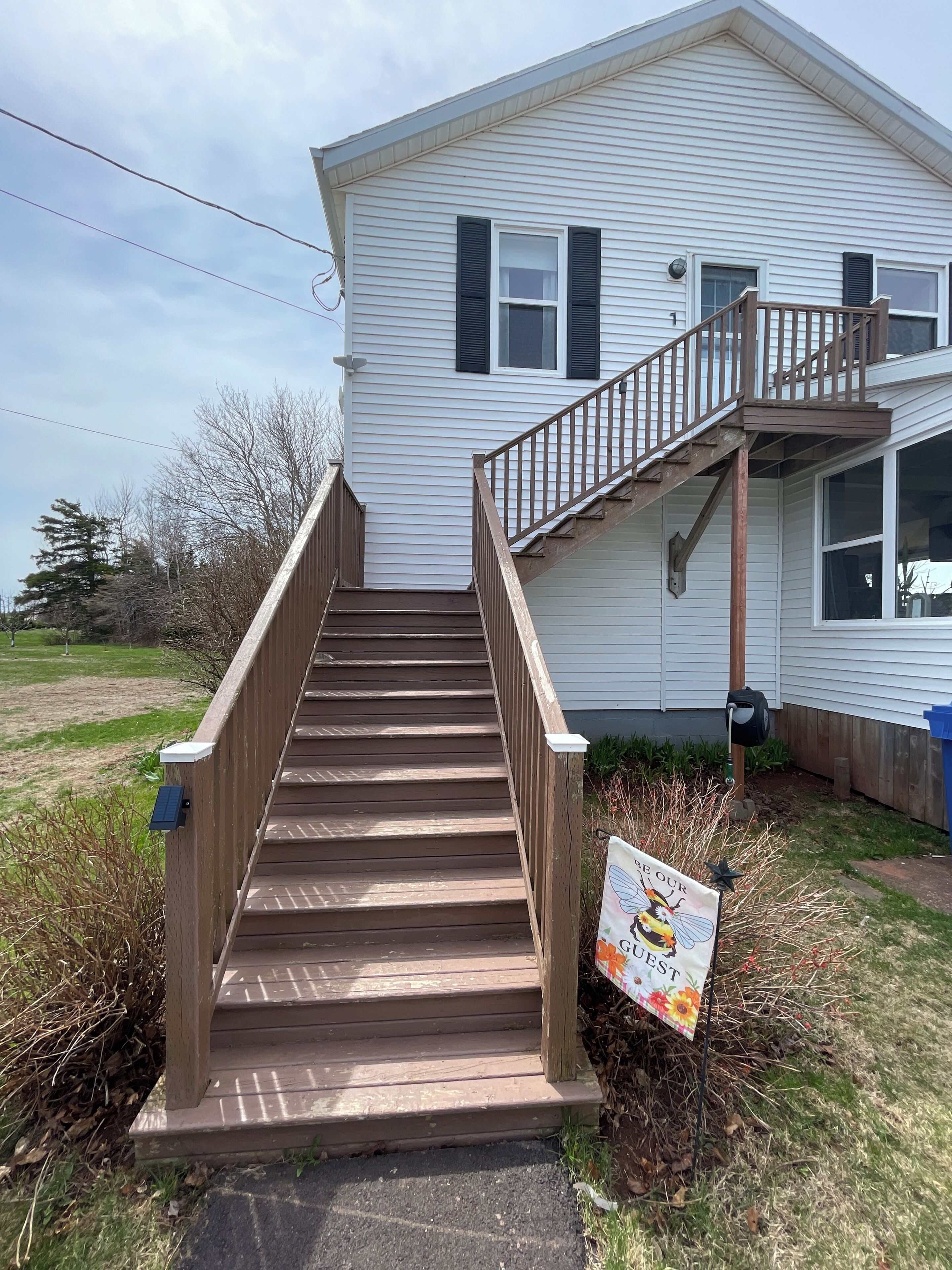 A wooden staircase leading to a white house with black shutters, a small deck, and a sign with a bee illustration.