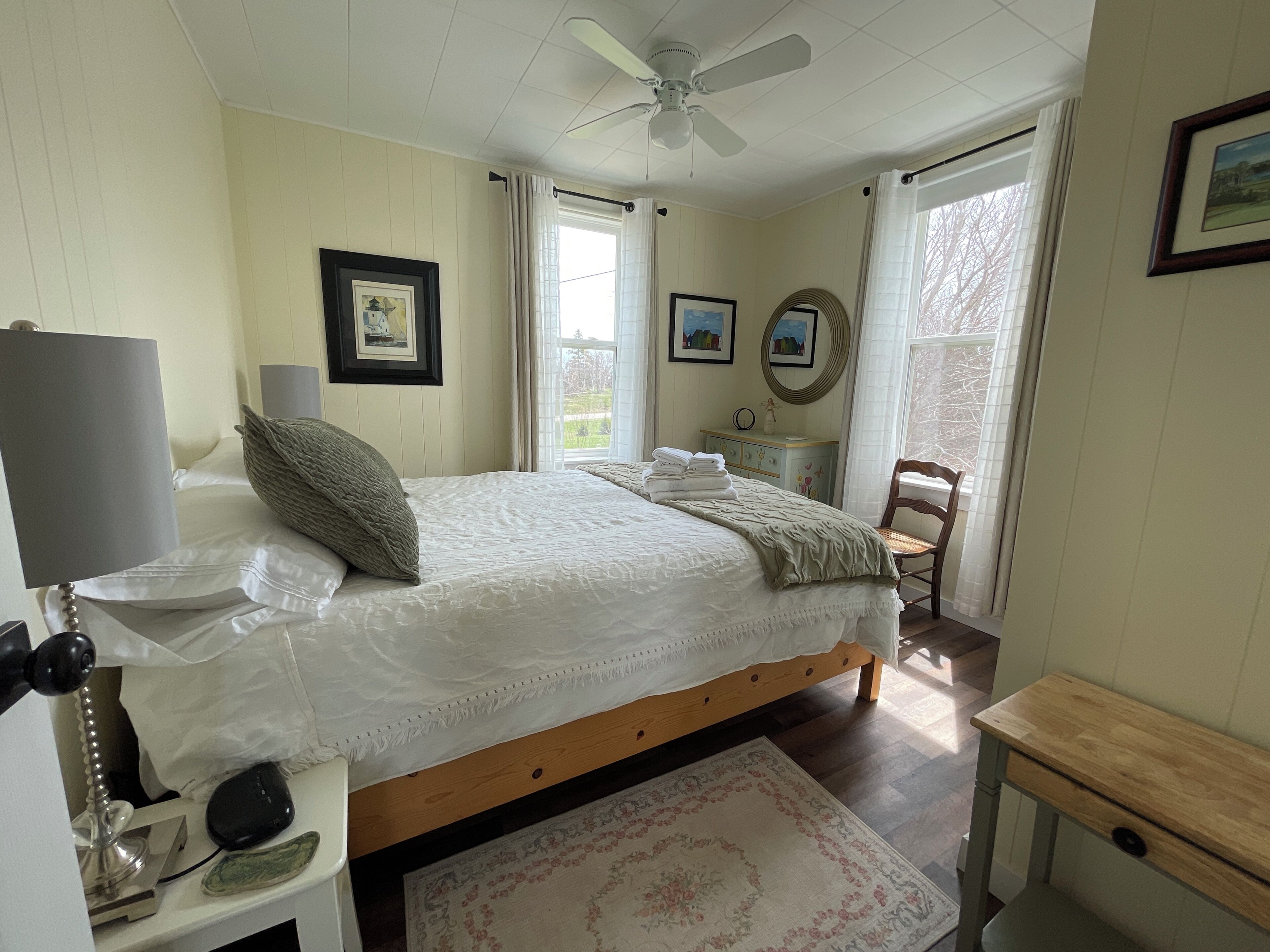 A bedroom with a bed, bedside table, lamp, chair, desk, and a ceiling fan.