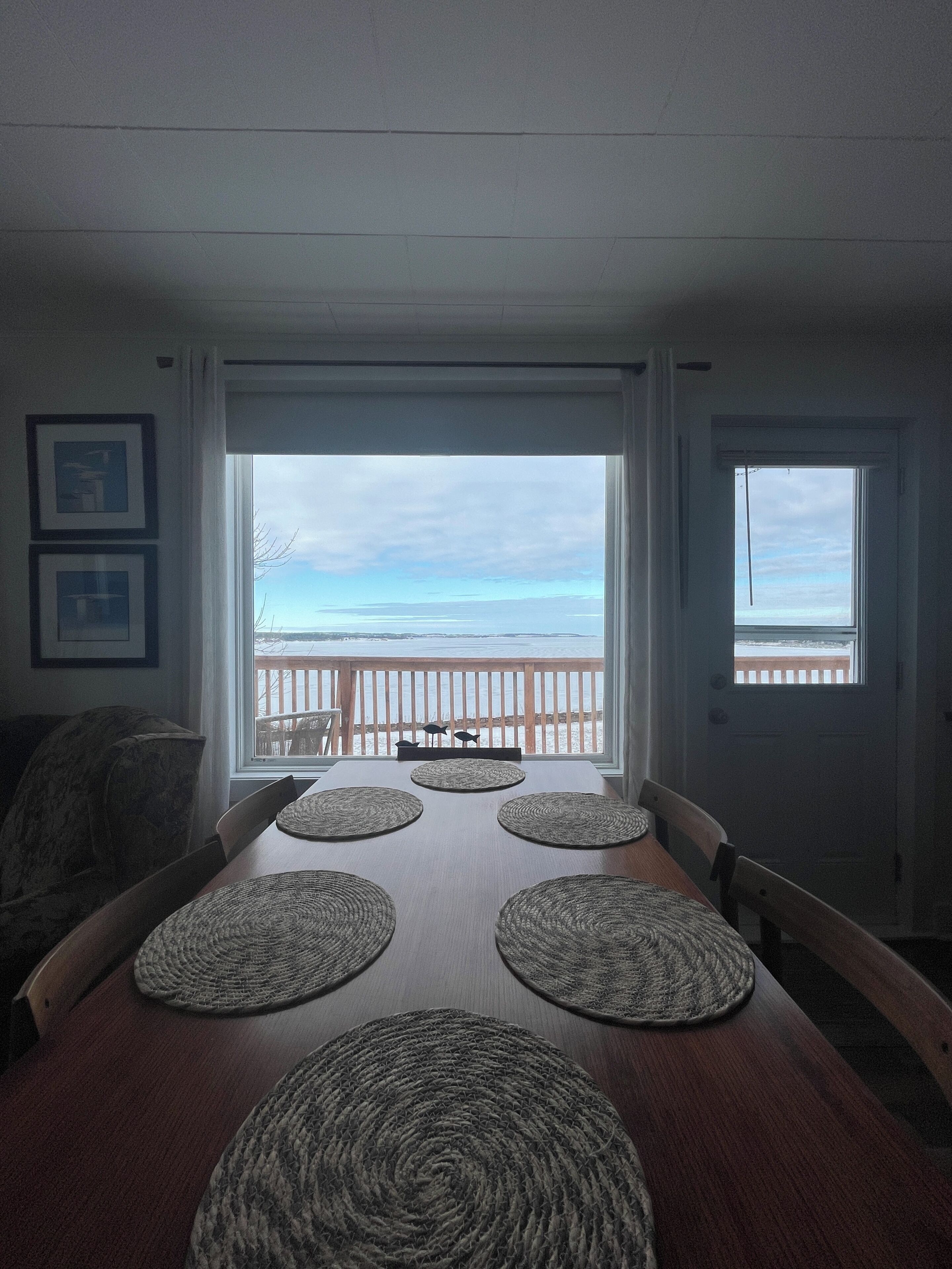 A dining area with a view of the ocean, a wooden table with placemats, and a balcony with a railing.