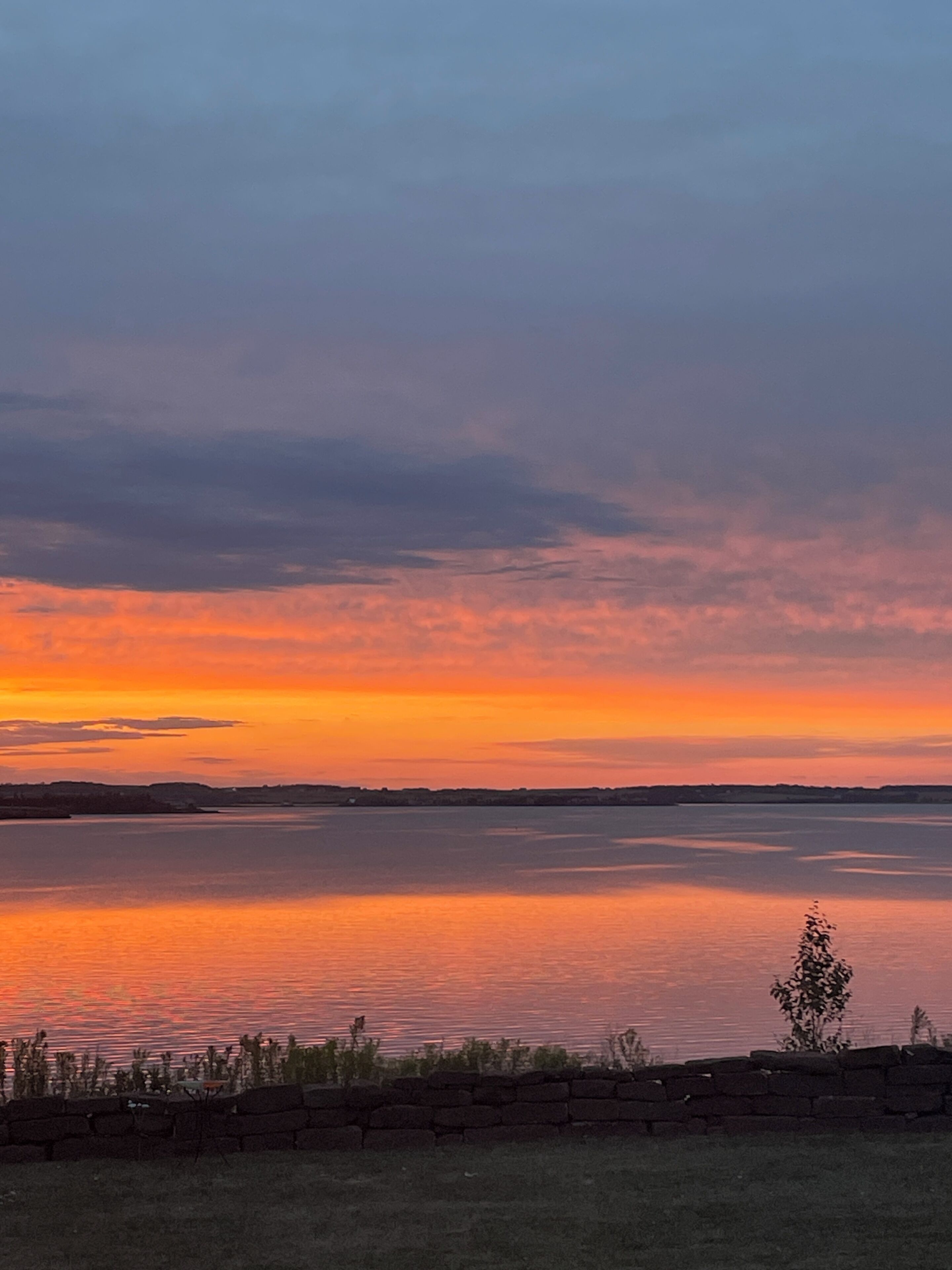 Een zonsondergang boven een rustig wateroppervlak, met een stenen muur en schaarse begroeiing op de voorgrond.
