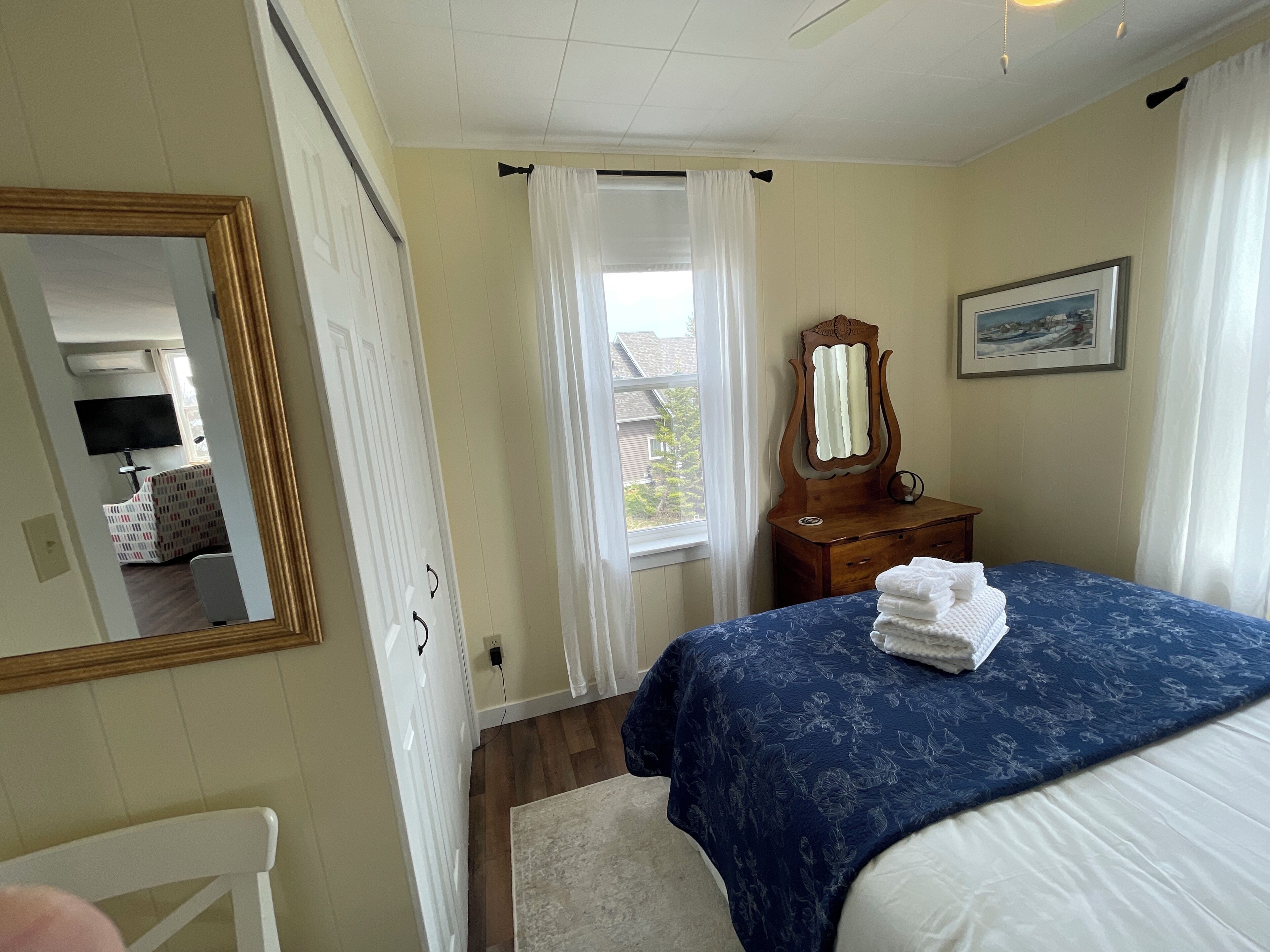 Een slaapkamer met een bed, een dressoir, een spiegel, een raam met gordijnen en een schilderij aan de muur.