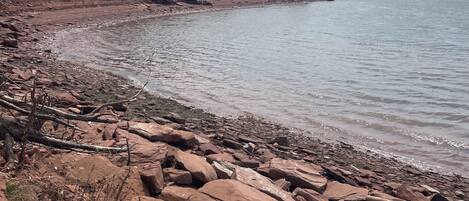 A rocky shoreline with a calm body of water, trees, and a distant house.