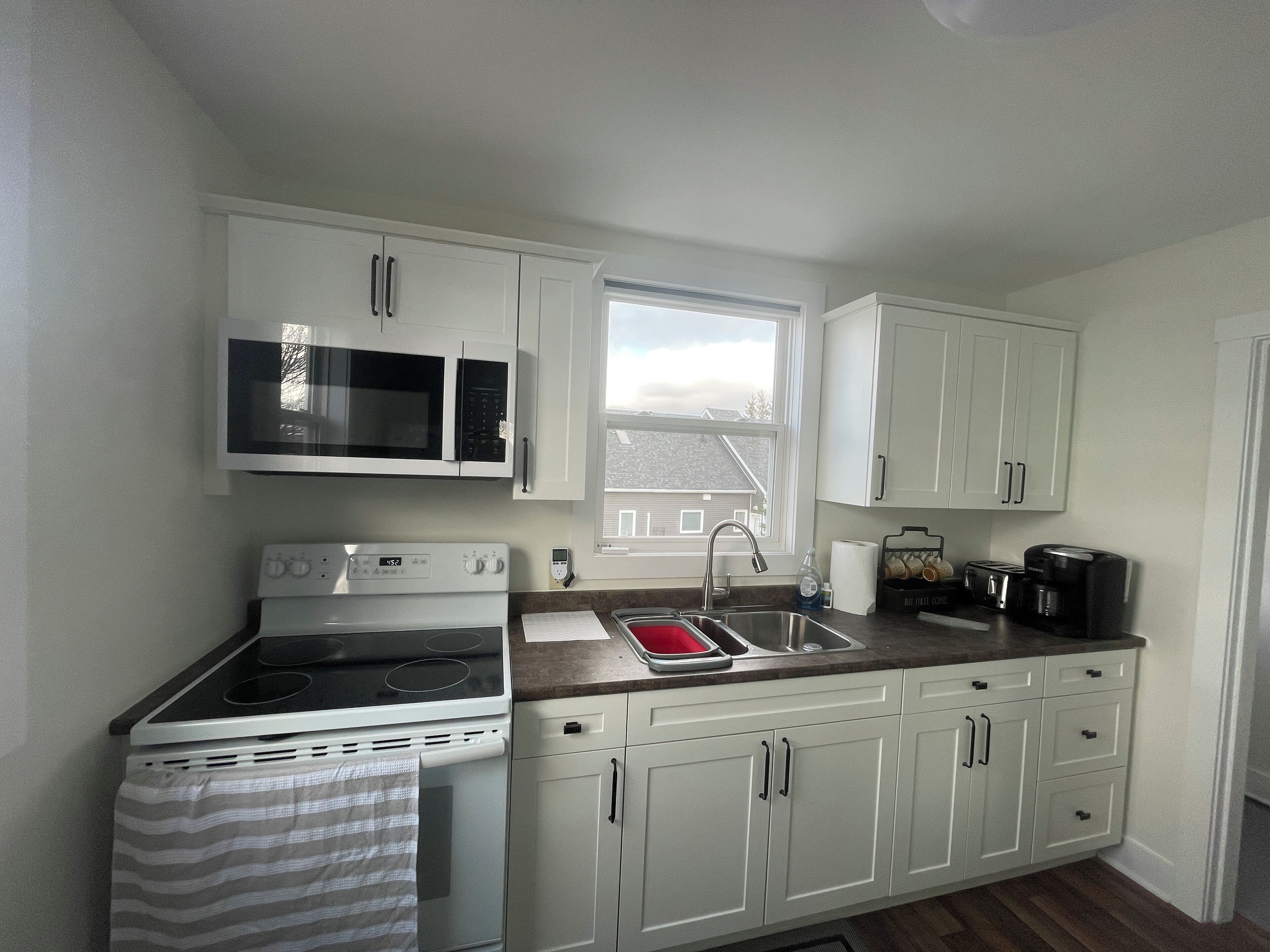 A kitchen with white cabinets, a microwave, stove, sink, and a window.