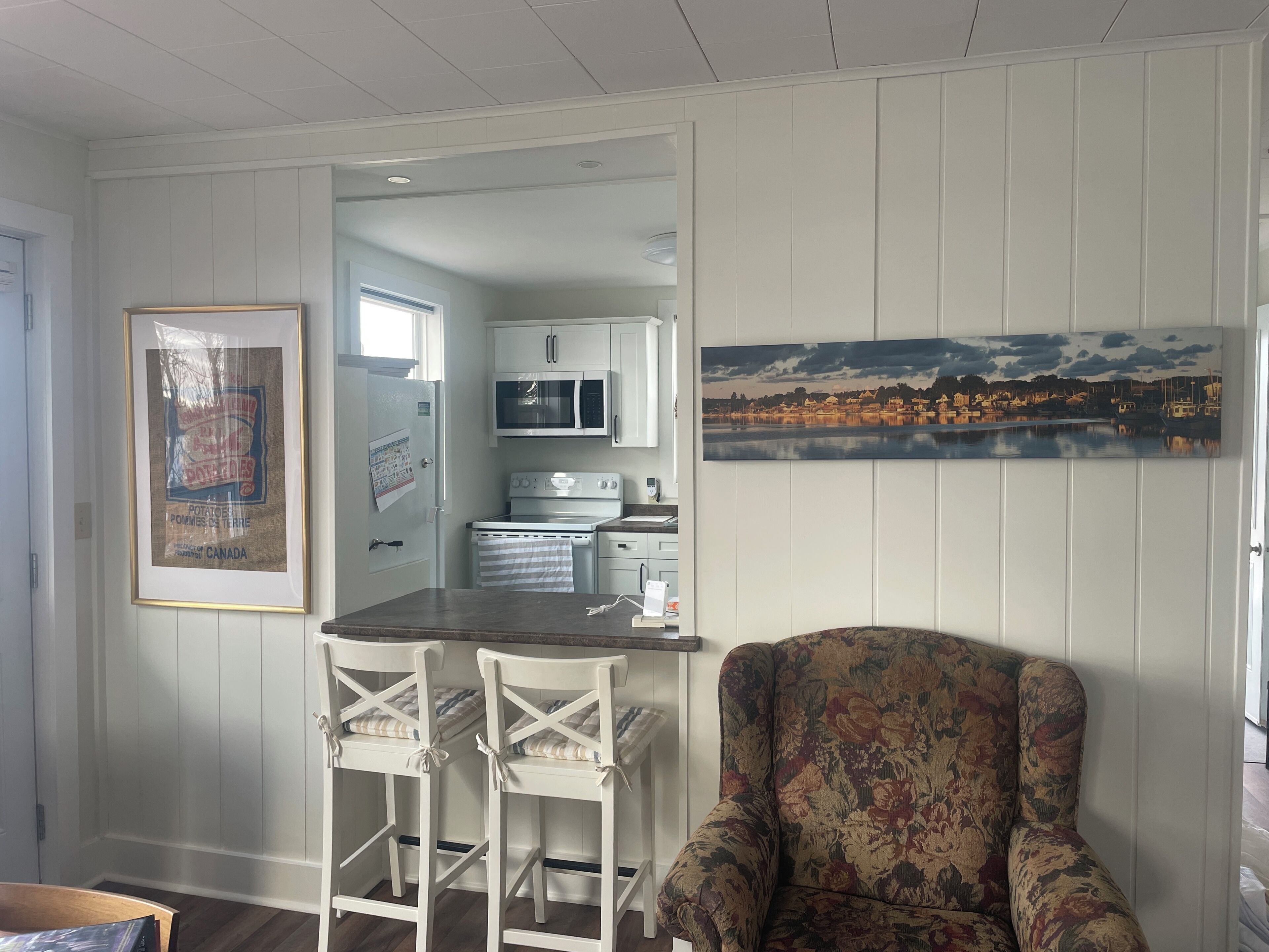 A kitchen with white cabinets, a microwave, and a stove. There is a framed picture on the wall and a chair with a floral pattern.
