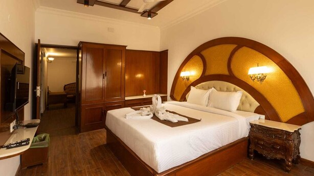 A hotel room with a large bed, wooden wardrobe, a television, and a ceiling fan. - WELBECK BY THE BEE HIVES (Ooty)