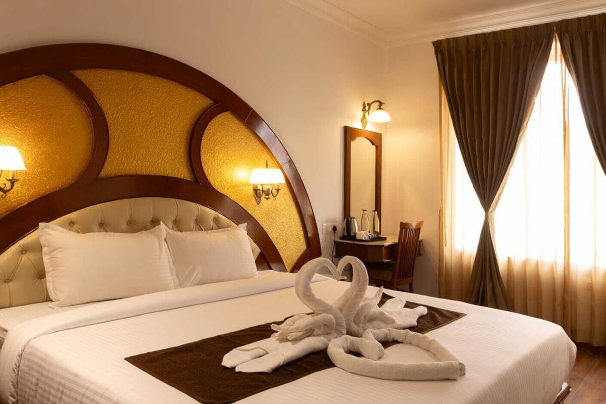 Une chambre d’hôtel avec un grand lit, une tête de lit agrémentée d’appliques murales, un miroir, une petite table avec une chaise et une fenêtre avec des rideaux.