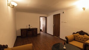 A room with a wooden floor, a dark round table, a sofa, a cabinet, and a door leading to another room. - WELBECK BY THE BEE HIVES (Ooty)