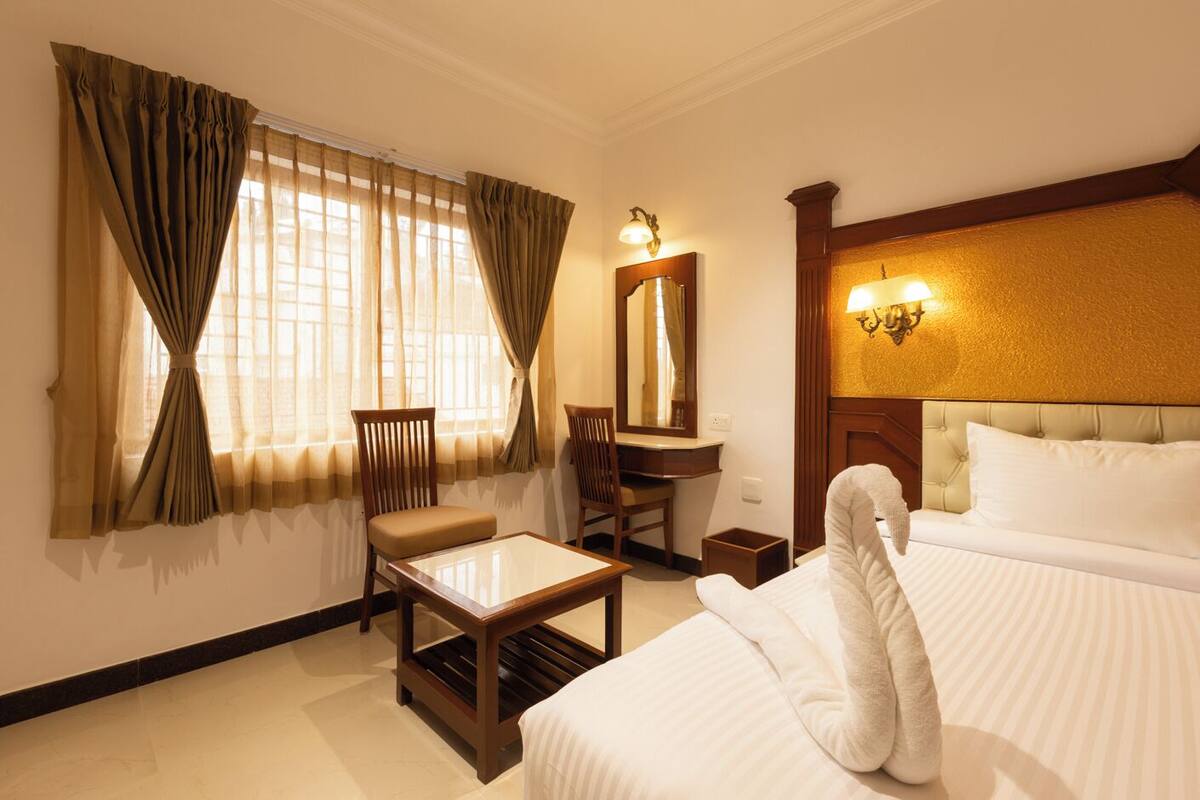 Une chambre d’hôtel avec un lit, un bureau, une chaise, une petite table, un miroir et une fenêtre avec des rideaux.